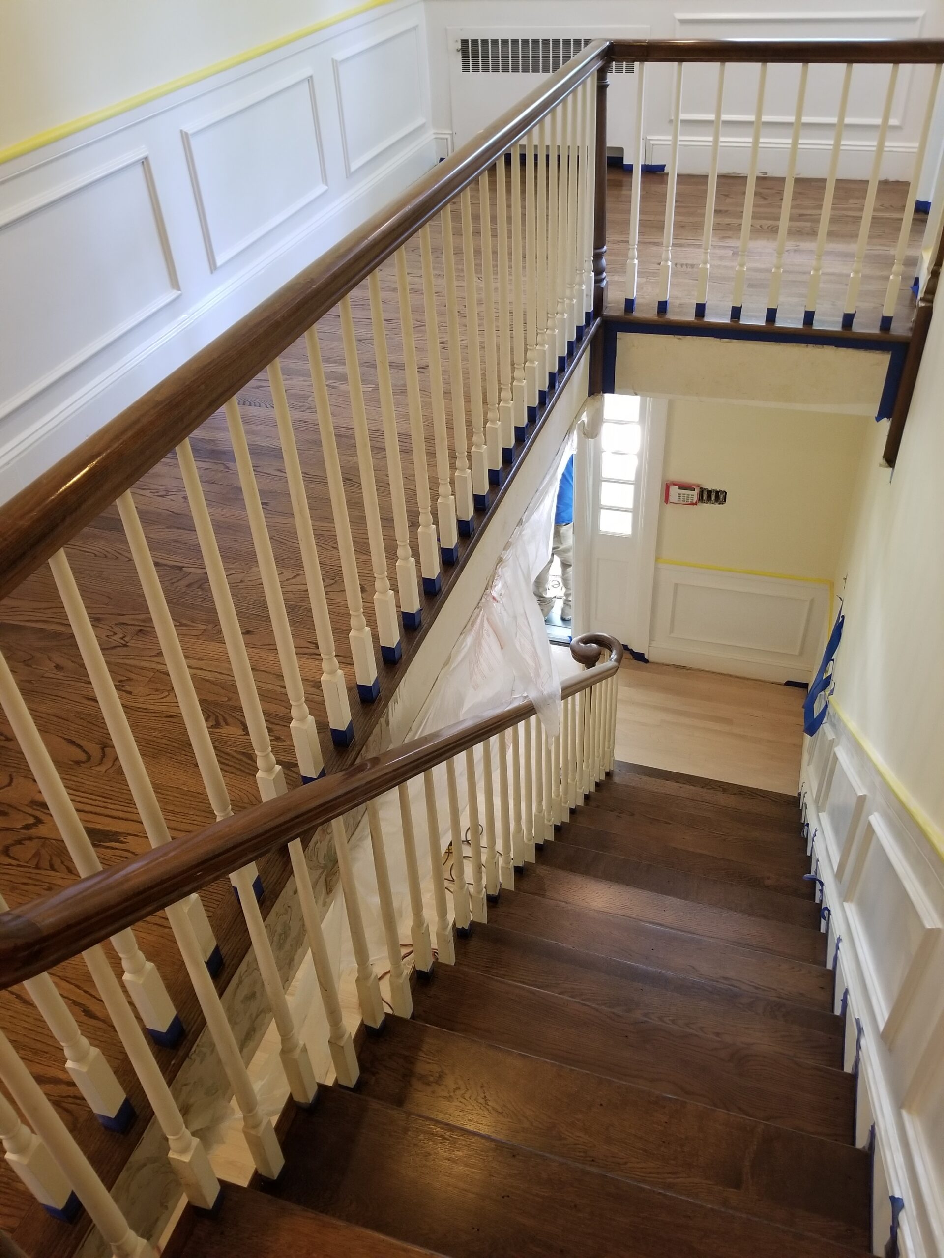Interior staircase with wooden steps and white railing, showing a hallway below. Painter’s tape and plastic sheet are visible, indicating ongoing painting or renovation work.