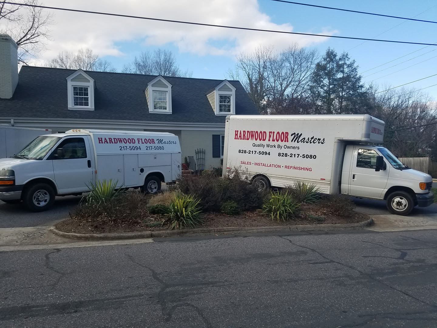 Two white service vehicles labeled Hardwood Floor Masters, a trusted flooring contractor, are parked in front of a gray house with dormer windows and a landscaped yard.