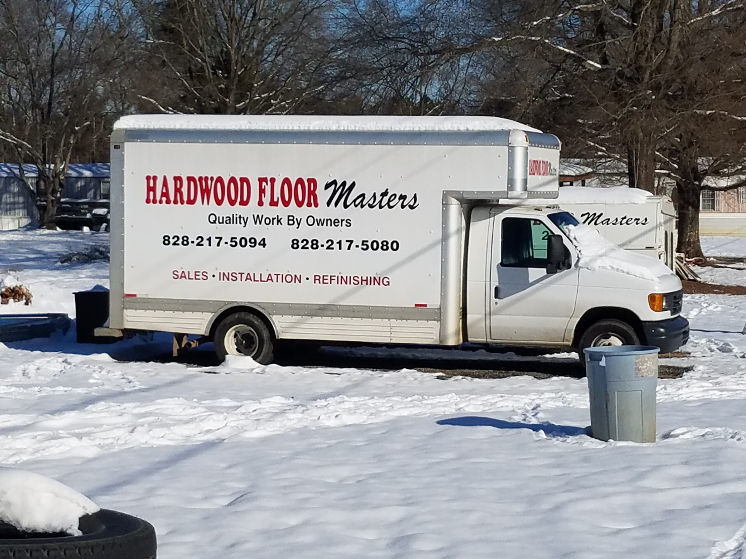 A white box truck labeled Hardwood Floor Masters, a trusted flooring contractor, is parked on snow-covered ground, with contact numbers and services listed on the side.