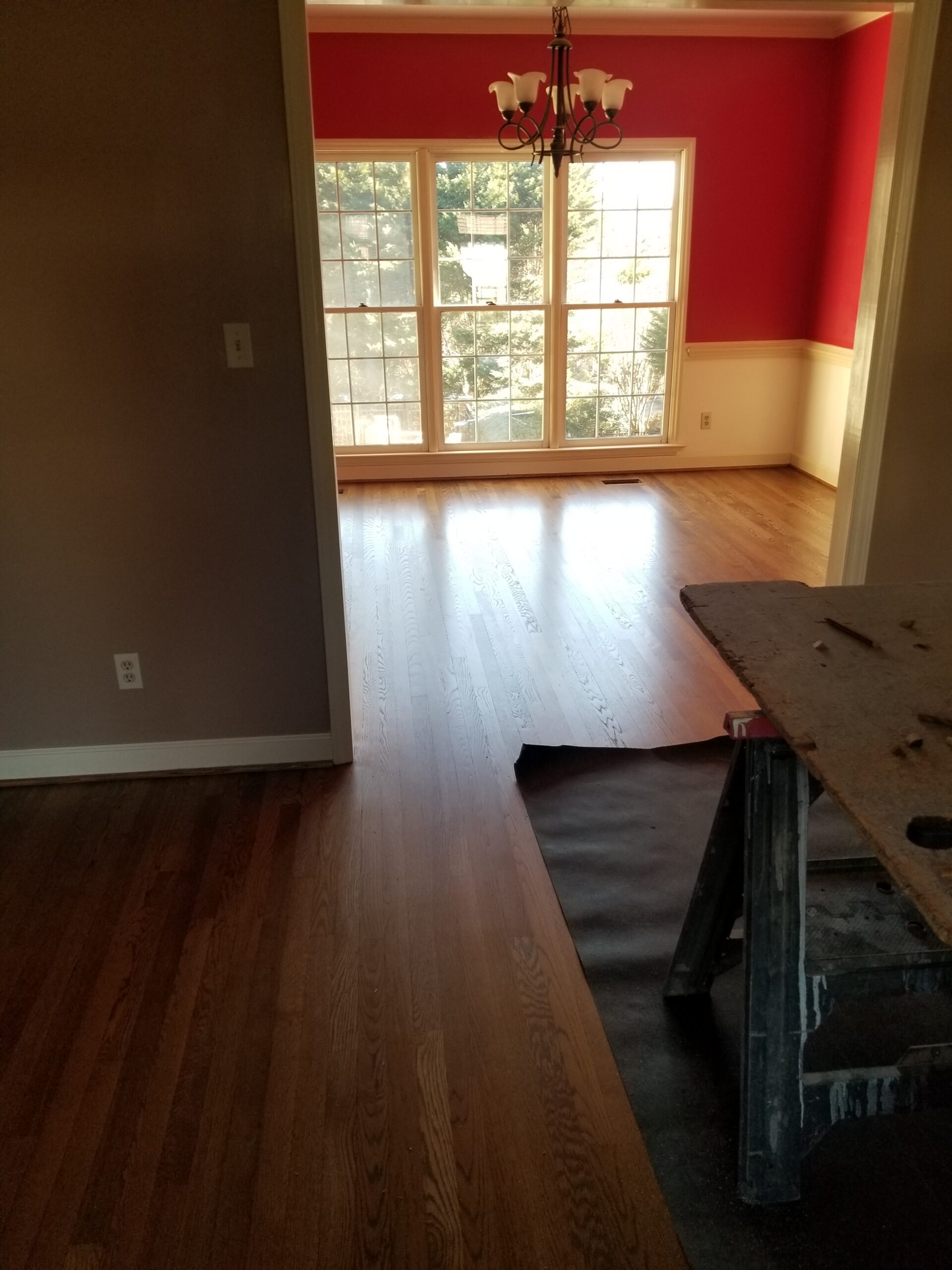 A flooring contractor installs hardwood floors in a room with a workbench in the foreground, while a bright dining area with red walls and large windows can be seen in the background.