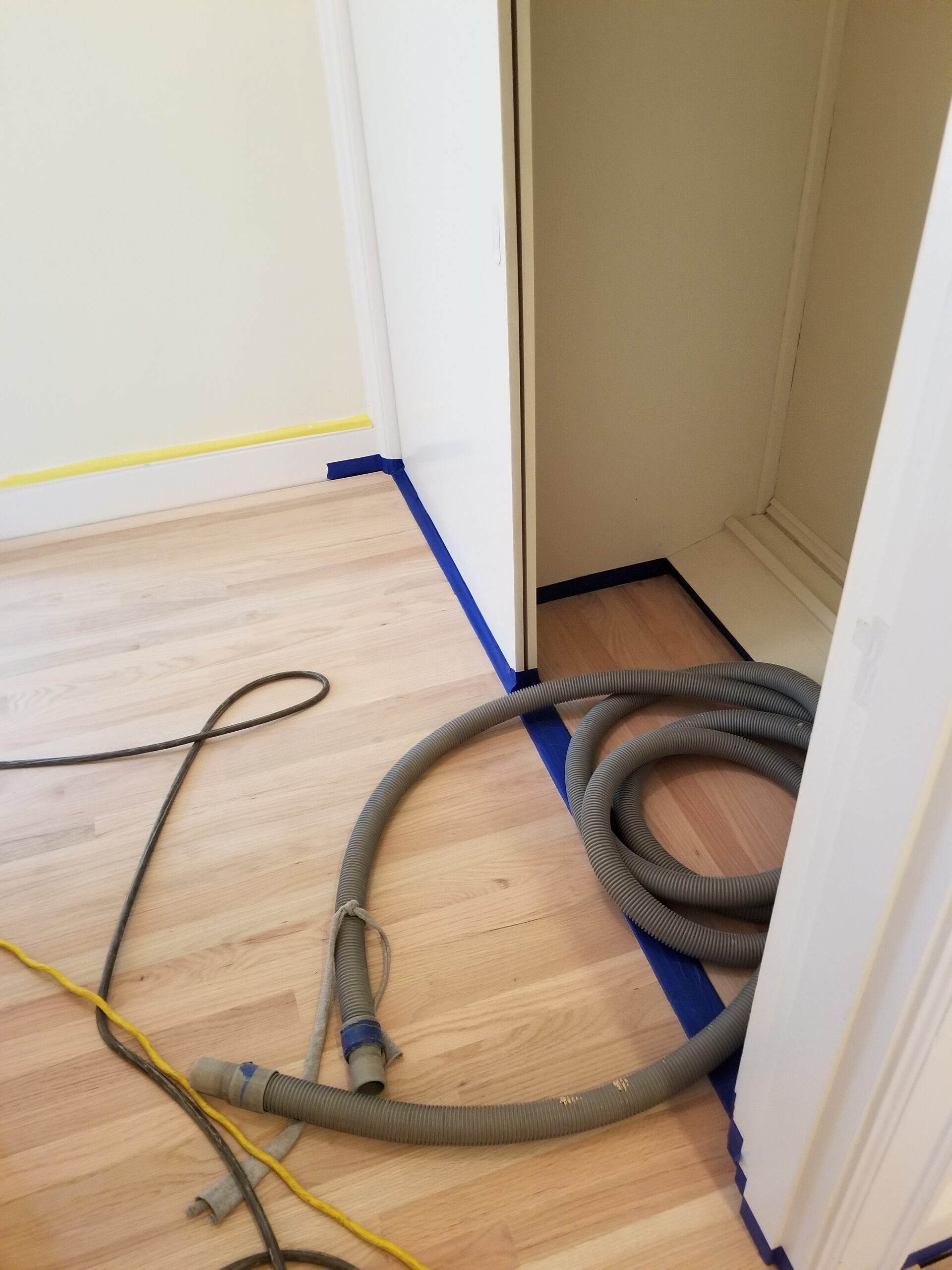 A flexible hose lies on a light wood floor near an open closet, with blue painter’s tape protecting the baseboards and closet edges—signs that a flooring contractor is preparing for installation or renovation work.