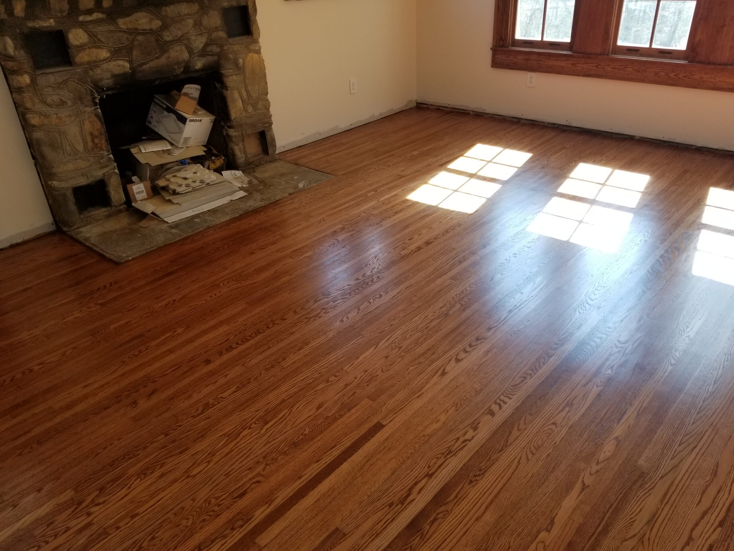 Empty room with a stone fireplace, unpacked items inside the hearth, and sunlight streaming through windows onto polished hardwood floors installed by a skilled flooring contractor.