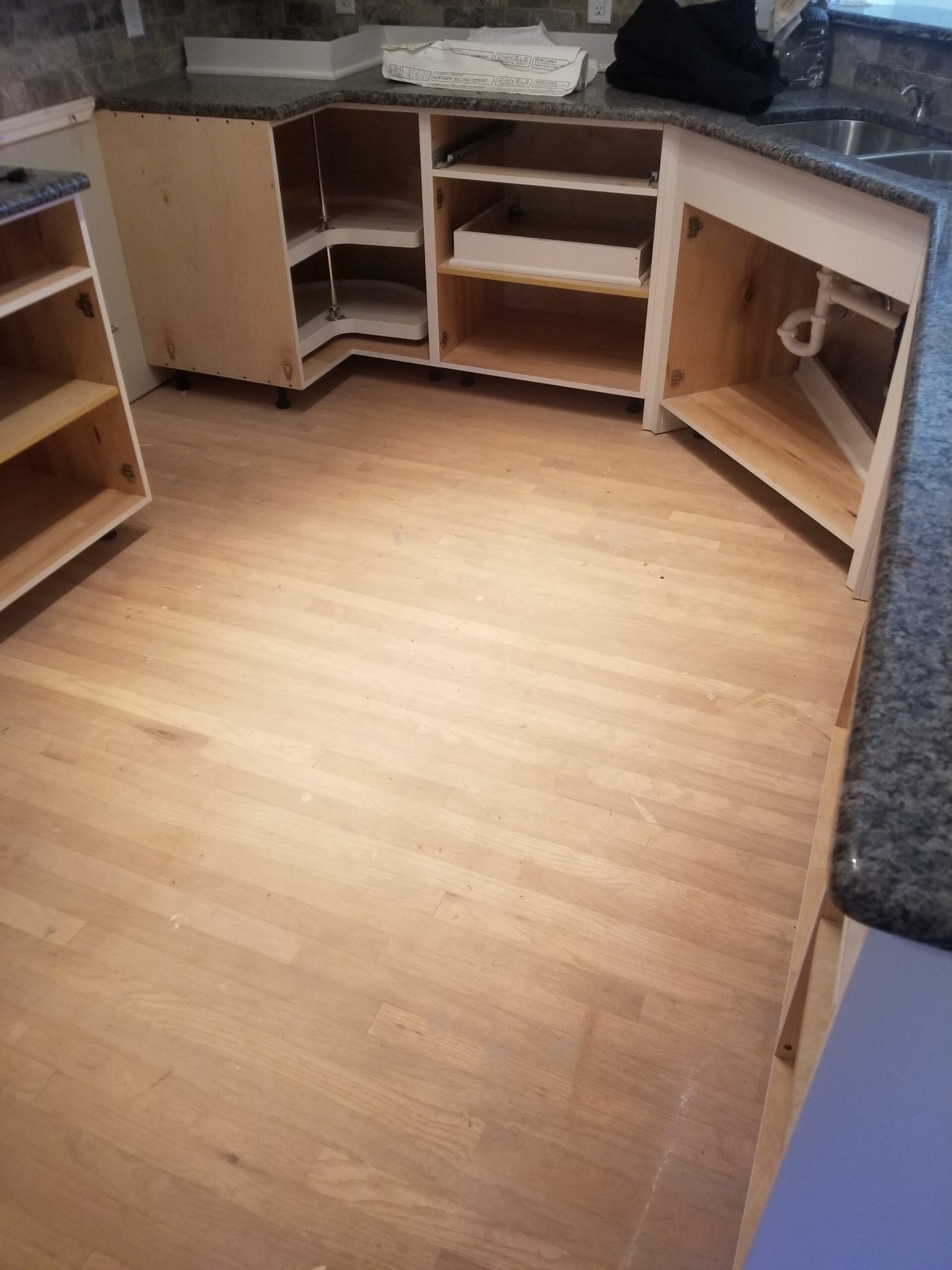 A kitchen with unfinished wooden cabinets, open doors, and no drawers or shelves installed, revealing exposed plumbing under the sink—ready for a flooring contractor to complete the space.