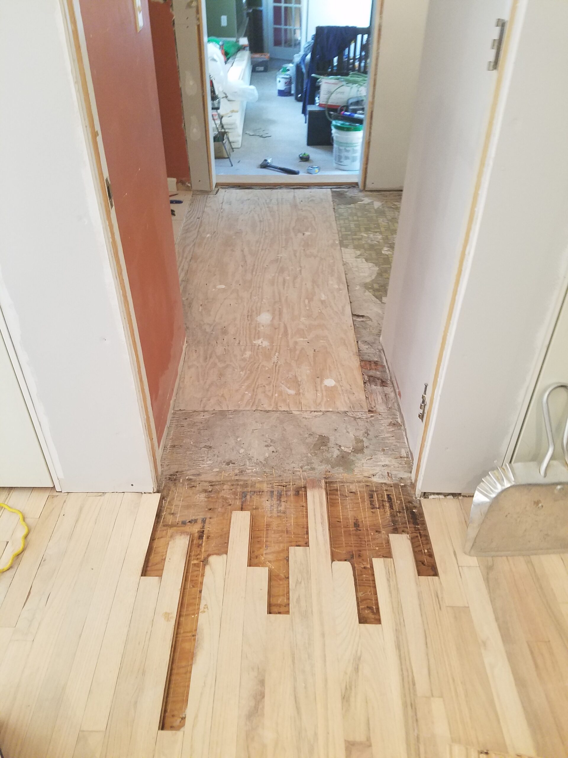 A hallway with partially installed hardwood flooring by a skilled flooring contractor, exposed plywood and subfloor, and construction materials visible in an adjoining room.