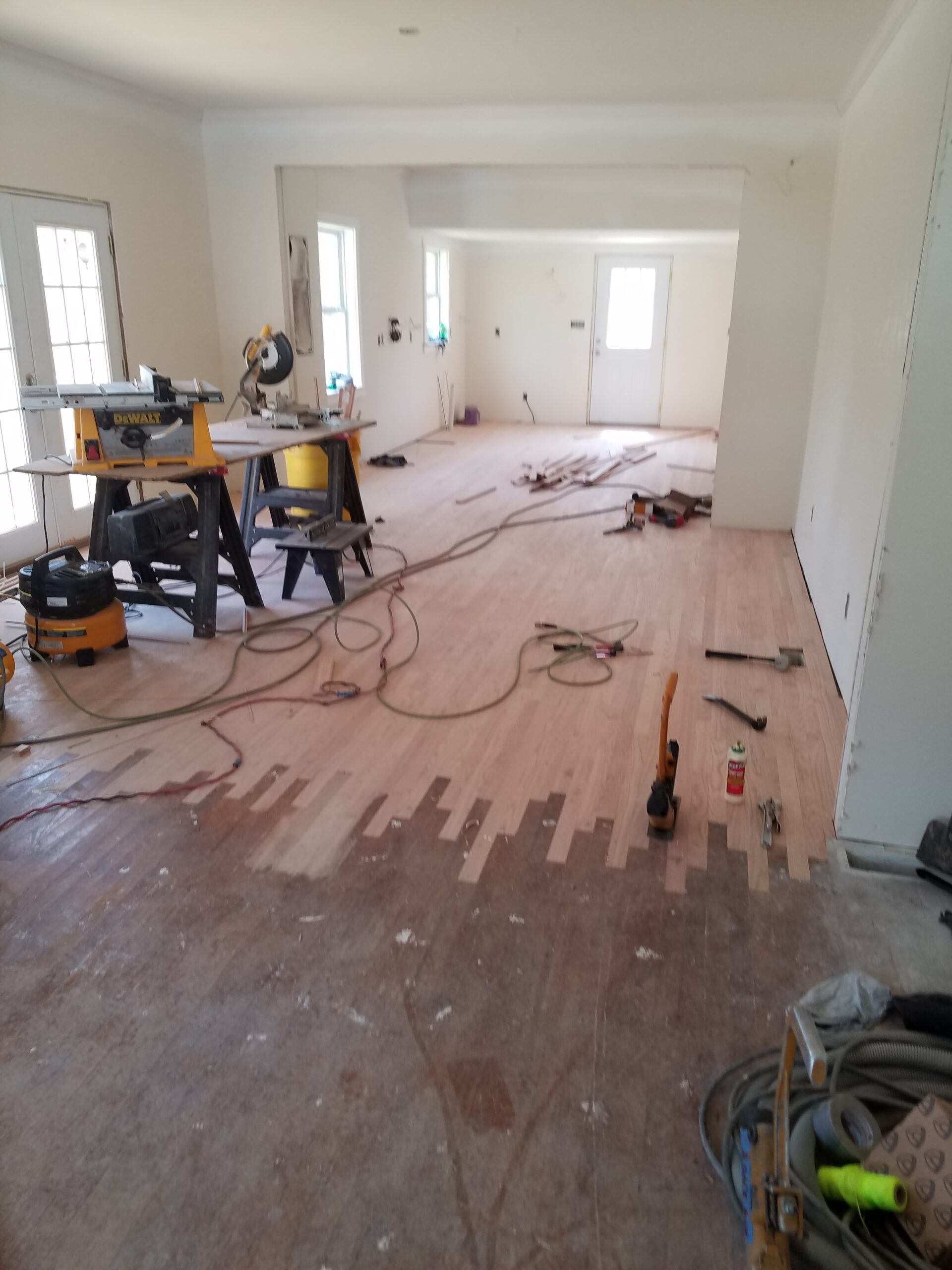 A room under renovation with new wooden flooring partially installed by a flooring contractor, tools and construction materials scattered around, and unfinished walls.