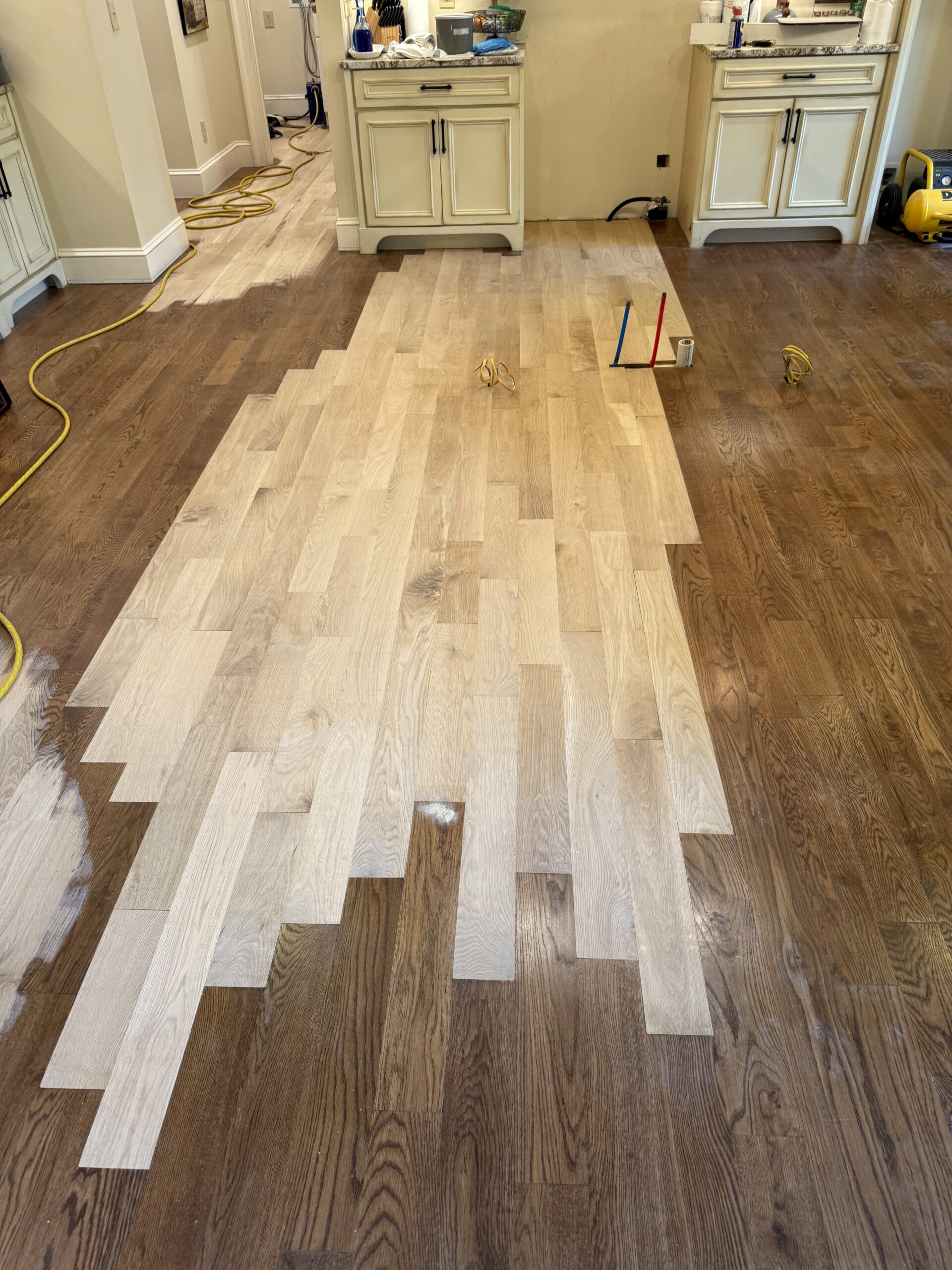 Partially refinished hardwood floor shows a clear contrast between sanded, unfinished wood and the darker, previously finished surface in a kitchen area, highlighting the skill of the flooring contractor. Tools and equipment are visible nearby.