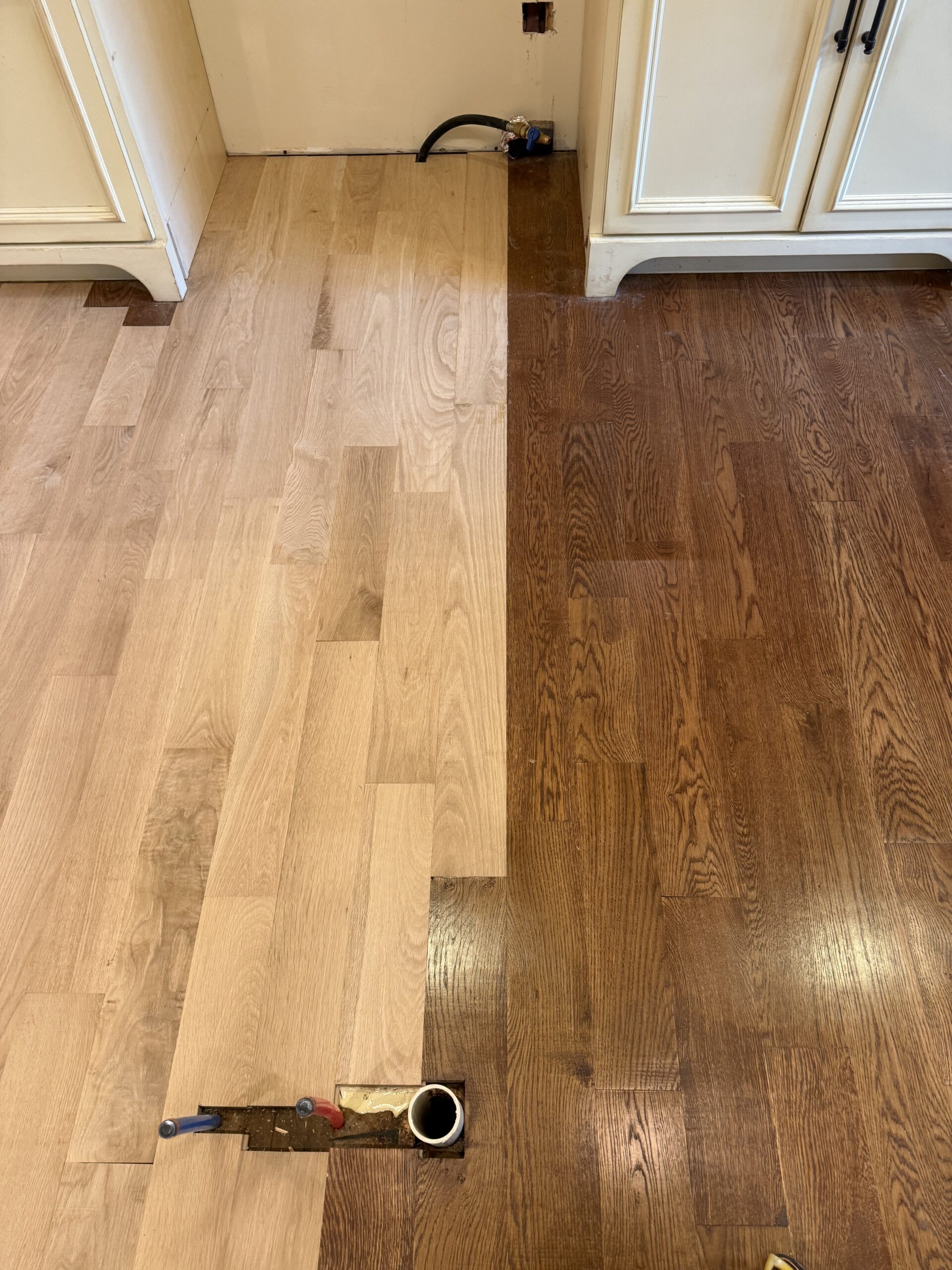 A hardwood floor, expertly worked on by a flooring contractor, is shown half unfinished on the left and half stained a rich dark brown on the right, with plumbing pipes visible in the lower part of the image.