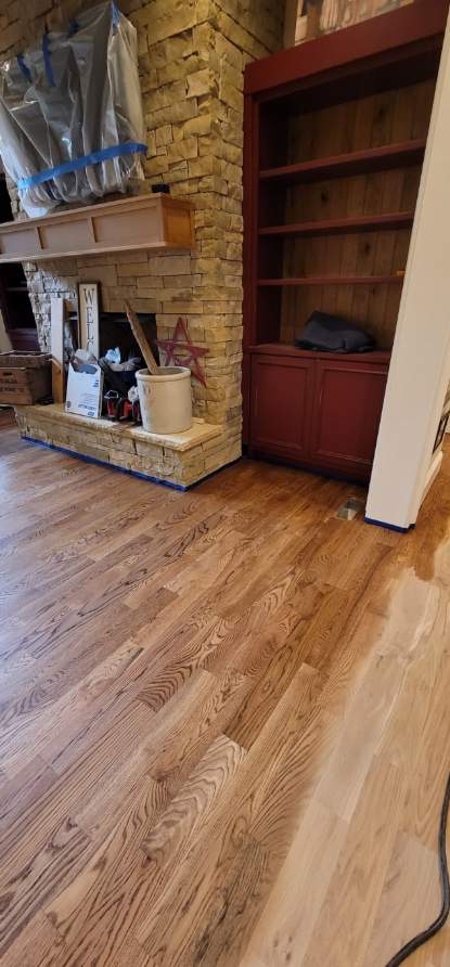 Living room with newly finished hardwood floors by a skilled flooring contractor, a stone fireplace with mantel, and an empty red bookshelf against the wall.
