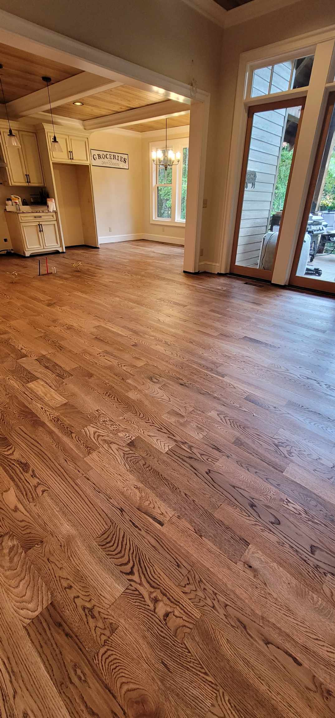 A spacious room with newly installed light brown hardwood floors by a skilled flooring contractor, adjacent to a kitchen and dining area with large windows and glass doors.